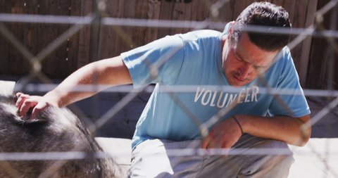 A Caucasian male volunteer wearing a blue uniform at an animal shelter, petting a rescued dog while feeding it on a sunny day in slow motion