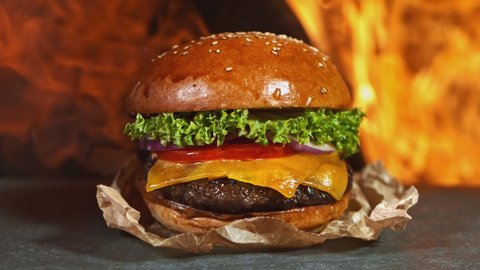 Tasty cheeseburger, lying on vintage wooden cutting board with fire flames in background. Super Slow motion filmed on cinema camera at 1000 fps