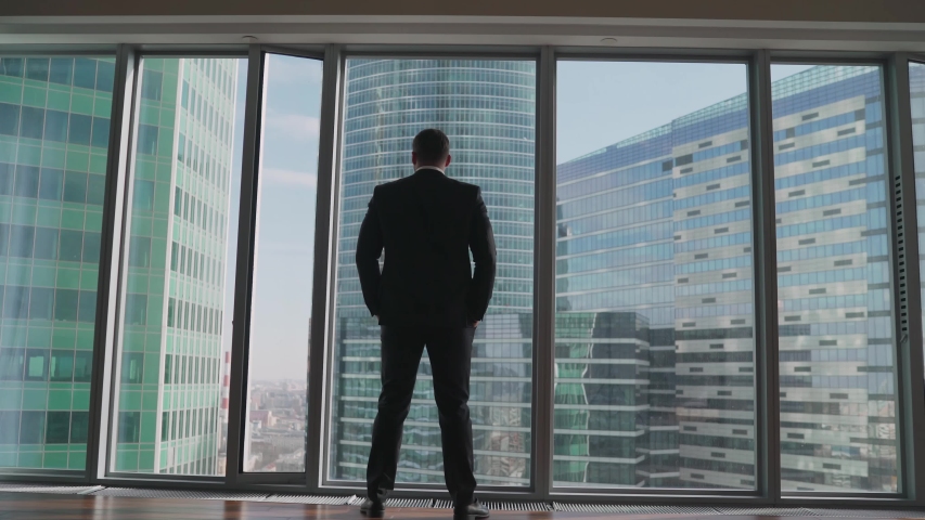 Successful businessman stands by the panoramic window. Back View of the Thoughtful Businessman wearing a Suit Standing in His Office, Hands in Pockets Looking out of the Window. Big City Business Royalty-Free Stock Footage #1053344834