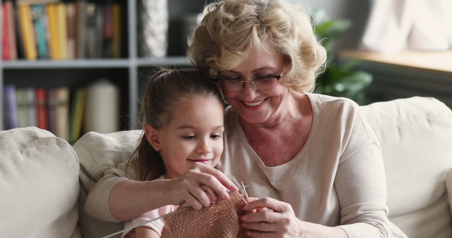 Happy elderly retired grandmother showing little cute granddaughter how to knit warm clothes with woolen threads in living room. Smiling different generations family enjoying hobby time indoors. | Shutterstock HD Video #1053348443