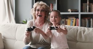 Happy pleasant elderly senior grandmother in glasses holding , playing online video games on tv with overjoyed little preschool child granddaughter, celebrating winning competition at home.