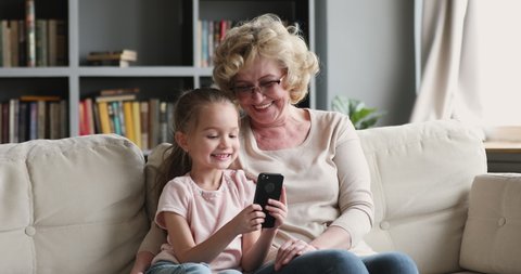 Adorable small kid girl holding cellphone, taking funny selfie shot with smiling mature grandma in glasses, relaxing together on couch. Little granddaughter playing mobile games with granny at home.
