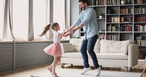 Full length happy millennial daddy inviting to dance small princess daughter at home. Smiling little preschool child girl holding father's hands, twisting to music together in modern living room.