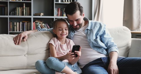 Happy cute small kid girl showing funny mobile app to smiling young father, resting together on comfortable couch in living room. Cheerful dad enjoying spending time with little child daughter.