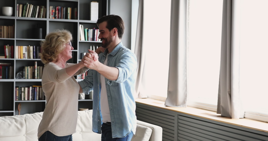 Happy young handsome man dancing waltz with smiling beautiful middle aged mother in living room. Excited senior older woman enjoying moving in pair to favorite slow music with grownup son at home. Royalty-Free Stock Footage #1053352166