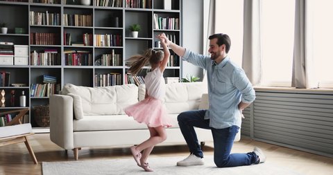 Full length smiling young daddy standing on one knee, twisting happy little preschool princess kid daughter, kissing hand after finishing dancing in modern living room, family hobby weekend pastime.