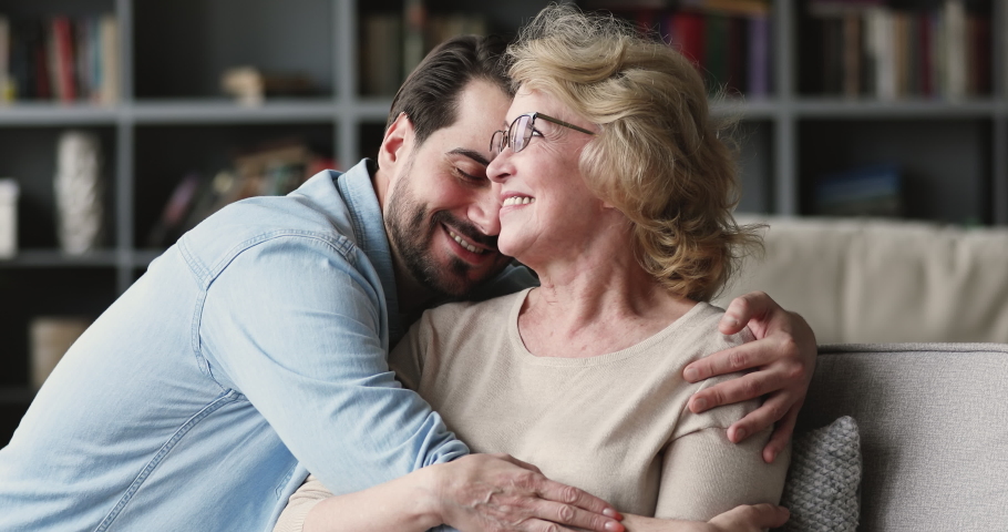 Happy young man visiting, coming to positive beautiful mature elderly mommy. Smiling millennial grownup son embracing, taking care of affectionate middle aged mother, relaxing on sofa at home. | Shutterstock HD Video #1053353576