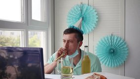 A man in a medical mask sits in front of a laptop and blows whistles, celebrates birthday with friends over the Internet. Celebrates birthday online, quarantine self-isolation.