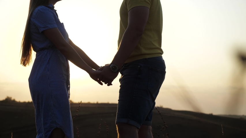 Beautiful young girl holds her boyfriend's hands and cuddles with him in a deserted field among the spikelets on a sunset background, focusing on her hands. Happy romantic relationship of cute couple. Royalty-Free Stock Footage #1053368822