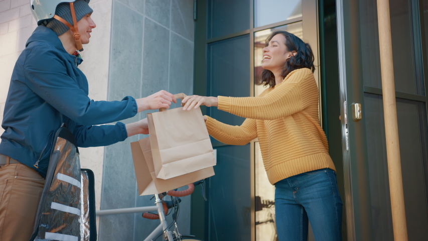 Happy Food Delivery Man Wearing Thermal Backpack on a Bike Delivers Restaurant Order to a Beautiful Female Customer. Courier Delivers Takeaway Lunch to Gorgeous Girl in Modern City District Office  Royalty-Free Stock Footage #1053374393