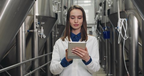 Portrait of beautiful female process engineer entering data on tablet at brewery. Young woman in white lab coat touching and typing digital display while standing and looking to camera.