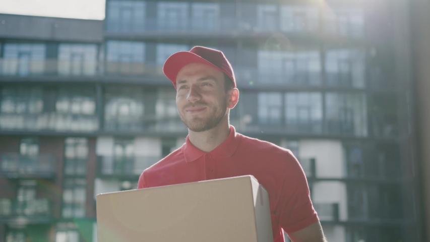 Cheerful handsome postman in red uniform delivering big cardboard box to beautiful customer young woman in modern business district. Home order delivery, transportation. | Shutterstock HD Video #1053377636