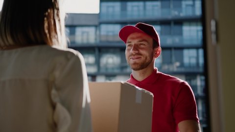 Cheerful handsome postman in red uniform delivering big cardboard box to beautiful customer young woman in modern business district. Home order delivery, transportation.