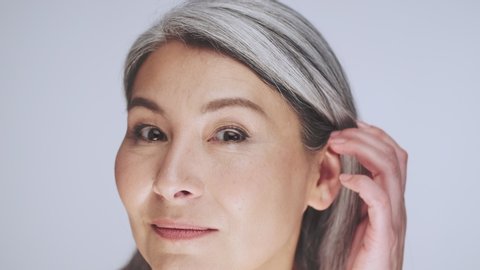 An attractive old mature half-naked woman with long gray hair is looking like to the mirror at her perfect skin and correcting hairstyle isolated over white background in studio