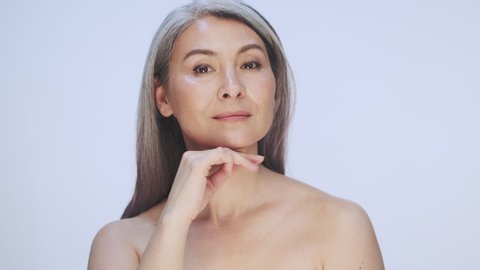 A gorgeous old mature half-naked woman with long gray hair is posing and touching her perfect skin isolated over white background in studio