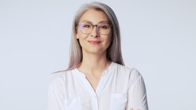 A pleased old mature woman with long gray hair wearing glasses is looking to the camera standing isolated over white background in studio