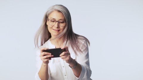 An emotional joyful old mature woman with long gray hair wearing glasses is playing games on her smartphone isolated over white background in studio