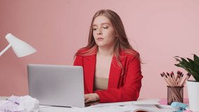 A serious tired young woman is working using her laptop computer sitting at the table isolated over pink background