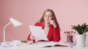 A focused young woman is reading paper documents something while sitting at the table isolated over pink background