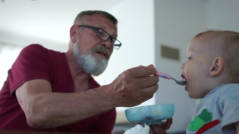 Grandfather in glasses and with a gray beard feeds from the spoon of his grandson. Happy family of grandfather and grandson