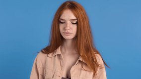 Red-haired girl in beige jacket. She coughing and putting on white mask while posing on blue studio background. Coronavirus pandemic. Close up