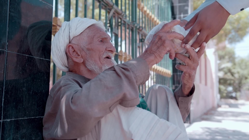 Poor old Pathan Afghan Muslim man, sitting on the footpath receives food from thoughtful social workers during the pandemic. Karachi, Pakistan. 22nd April 2020. | Shutterstock HD Video #1053396764