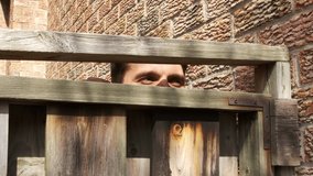 High quality close-up 4k slow motion video of a suspicious man peeking out wooden fence, man watching neighbours. Handsome spy.