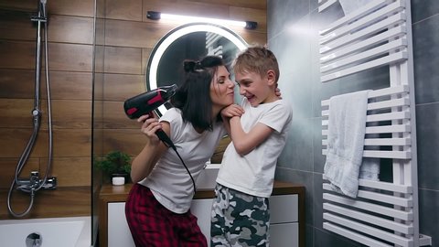 Pretty smiling carefree young woman kissing,tickling her funny handsome son during drying his hair with hair dryer in the bathroom