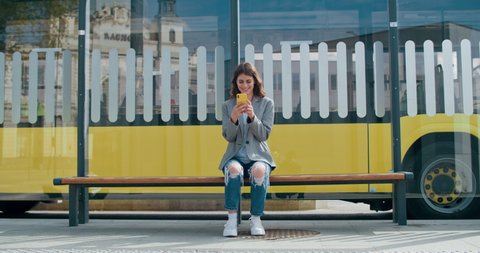 Cheerful woman wearing casual clothes browsing internet or chatting in social networks. Pretty girl using smartphone while sitting at public transport stop. Bus riding at background