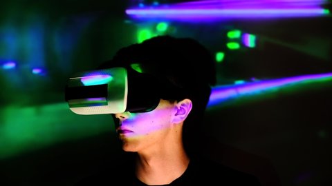 Young guy looking around surprised. Man watches and using VR-helmet virtual reality night at home in front of screen. Portrait men at exhibition, club party with dynamic projector illumination.