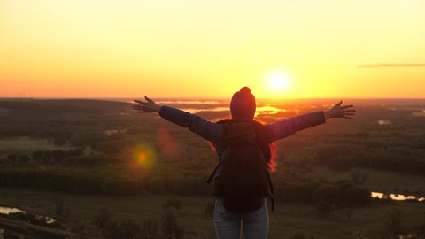 young woman traveler with backpack comes to the edge of a mountain peak sunrise, raises his hands in the air and enjoys victory, beautiful sun and landscape. free girl tourist travels in nature alone