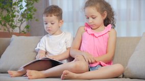 Cute children,sister with a younger brother, looking at a book magazine textbook dictionary tablet read fairy tales, kids are reading books sitting on the couch,preschool and school education.