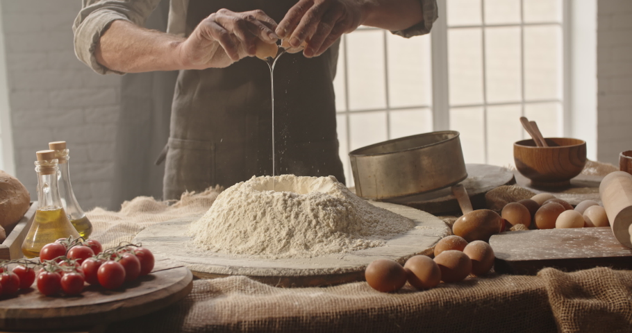 Experienced mature chef cracking an egg into flour to make bread according to traditional recipe. Old man baking at home, enjoying hobby - closeup shot 4k footage | Shutterstock HD Video #1053414959