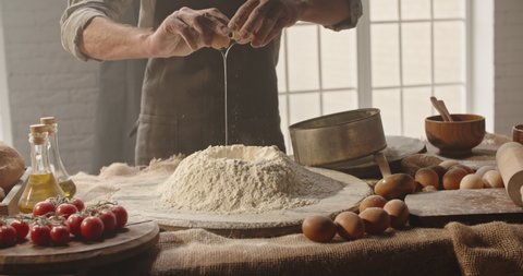 Experienced mature chef cracking an egg into flour to make bread according to traditional recipe. Old man baking at home, enjoying hobby - closeup shot 4k footage