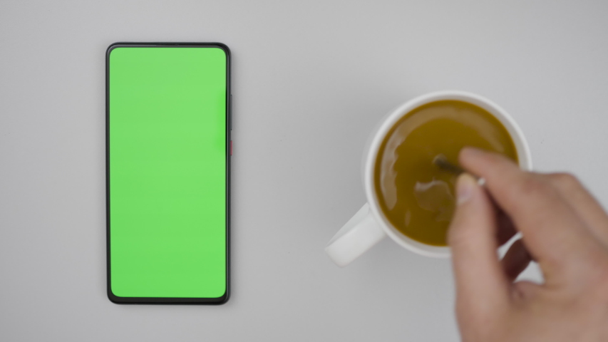 Man Hand Cup of Hot Coffee and Using Smartphone Watching Green Screen Top View. Smartphone with Green Mock-up Screen Business Concept. Person Hand Stirring Coffee with Spoon on Table. Slow Motion. | Shutterstock HD Video #1053420053