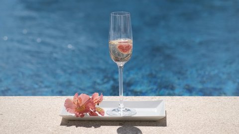 Close up of glass with champagne or prosecco with bubbles on swimming pool or sea background. Sparkling white wine on tray with flower. Raspberry falling in glass with cold champagne, luxury drink. 4K