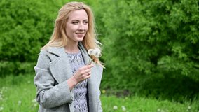 Video of a young caucasian pretty woman blowing on a flower in a park outdoors. Model is wearing a coat in spring.