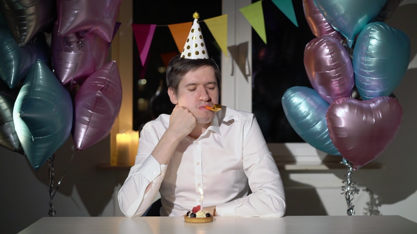 Young man celebrating birthday alone. Sad bored and tired male bday with party cake and blowing candle, balloons