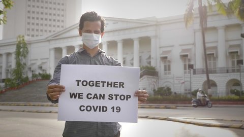 A young man wearing face mask standing and holding a placard with message 'Together we can stop COVID 19' next to Asiatic library on empty road during city lockdown amid coronavirus epidemic