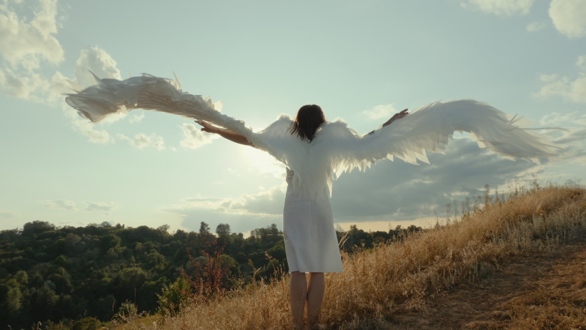 Angel with outstretched wings. Woman in a white dress raises angel wings up, looks at the sunny day, slow-motion shot