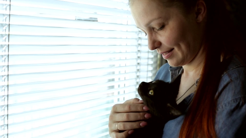a beautiful woman stands near the window and holds a black kitten in her arms, Pets