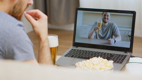 Back view of happy man on video call with his friend drinking beer during coronavirus isolation.