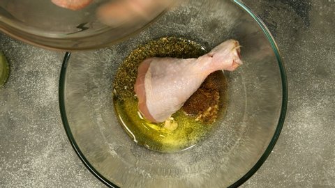 Chef prepares marinade and stirs raw chicken legs for barbecue or frying in deep glass bowl. Olive oil, spices, sauce are mixed with chicken drumsticks. Close-up.