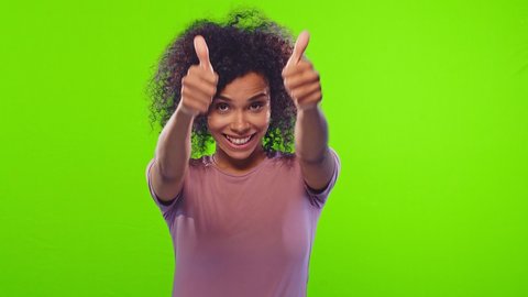 Charming cheerful beautiful african american positive woman showing thumbs up, gazes happy at camera and enjoying posing on green screen background. Positive feedback, body language concept