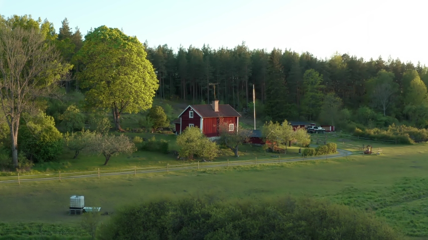 Swedish house in aerial drone shot. Small red typical wooden cabine home surrounded by forest and farmland fields with yellow canola rapeseed flowers. Summer evening light in Sweden countryside Royalty-Free Stock Footage #1053433805