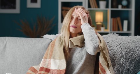 Unhappy woman with blond hair sitting on couch in warm blanket and suffering from headache. Mature female with blond hair coughing and sneezing while staying at home.