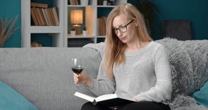 Beautiful woman in glasses and domestic outfit relaxing on grey couch and drinking red wine while reading interesting book. Concept of leisure time and enjoyment