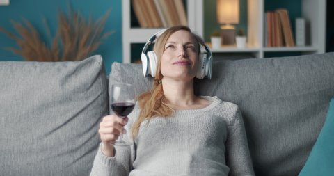 Portrait of beautiful woman listening music in wireless headphones while relaxing on grey couch with glass of red wine. Concept of leisure time and enjoyment