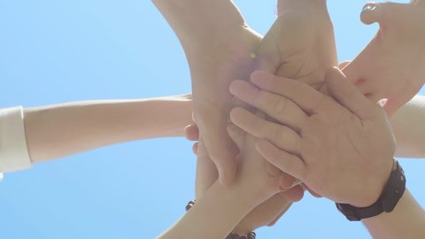 Vertical bottom view of people putting their hands together over blue sky background. Friends with stack of hands showing unity and teamwork. Team building, friendship and togetherness concept