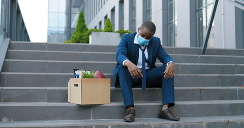 Fired African American male office worker in medical mask sitting on stairs in depression with box of stuff. Unemployed businessman lost his business. Anxious concept. Workless man in despair.